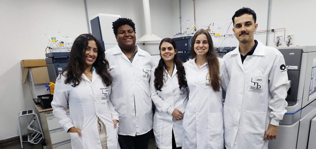 Cinco pesquisadores, três mulheres e dois homens, posam sorridentes lado a lado em um ambiente de laboratório. Todos vestem jalecos brancos com um logotipo bordado no bolso superior esquerdo. O grupo está posicionado à frente de equipamentos científicos complexos, que incluem máquinas de análise de grande porte com tubulações e fiações visíveis ao fundo, sugerindo um ambiente de pesquisa tecnológica ou bioquímica.