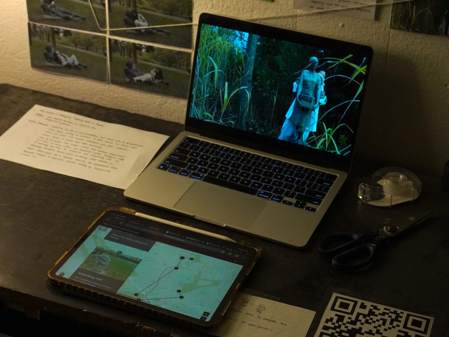 Installation view: A dimly lit desk features an open laptop playing a film scene of a person walking through tall grass with a backpack. Next to it, a tablet displays a digital map with marked locations. The desk is scattered with papers, a pair of scissors, tape, and a QR code, while printed film stills are pinned to the wall in the background.