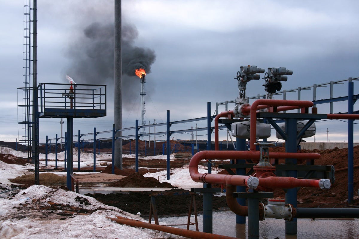 Methane gas being 'flared' at a fossil fuel production site. In the foreground are a complex series of pipes, valves, and other apparatus; dirty half-melted snow surrounds the site. Methane gas being 'flared' at a fossil fuel production site. In the foreground are a complex series of pipes, valves, and other apparatus; dirty half-melted snow surrounds the site.