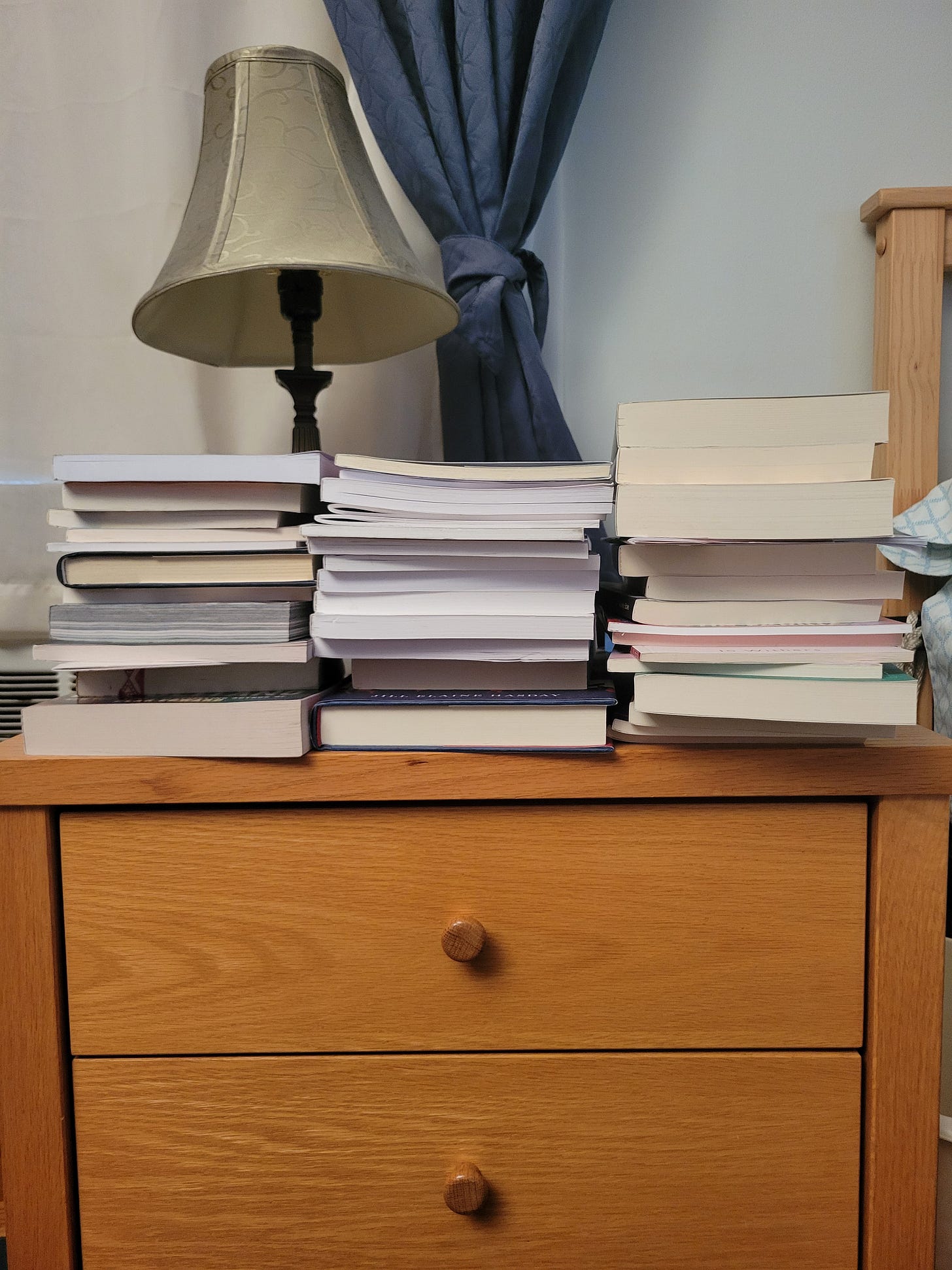 nightstand with three stacks of books and lamp in background, curtain behind the lamp and bedpost visible on the right, shades on the left