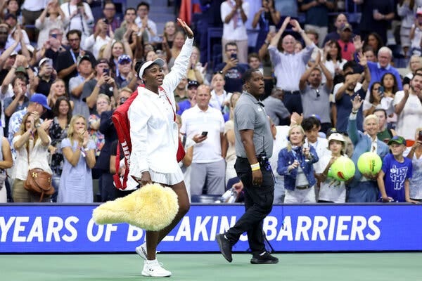 Venus Williams, dressed in white, holds a yellow-ish fuzzy racket bag while waving to fans. 