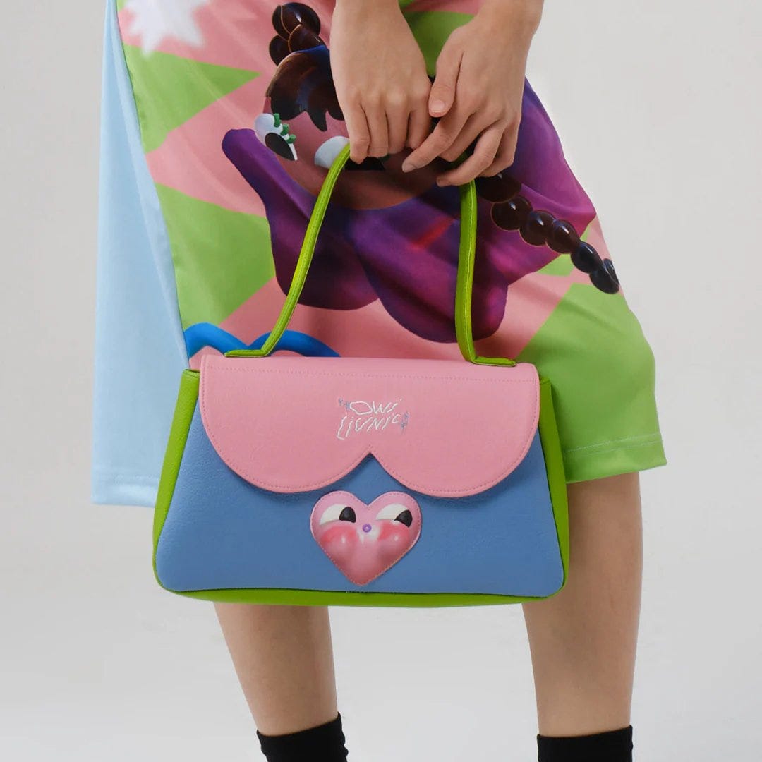Close-cropped photo of a person in a bright, cartoon-print skirt holding a small pastel handbag: pink scalloped flap, sky-blue body, lime-green trim and handles, plus a glossy pink heart with big eyes on the front panel. Close-cropped photo of a person in a bright, cartoon-print skirt holding a small pastel handbag: pink scalloped flap, sky-blue body, lime-green trim and handles, plus a glossy pink heart with big eyes on the front panel.