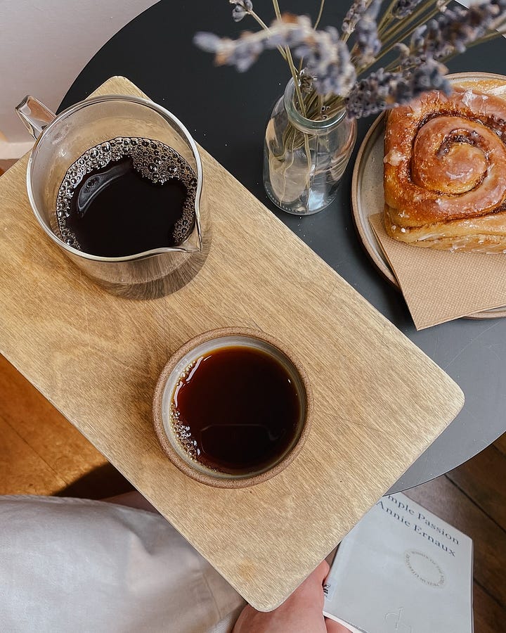 Image 1: Shopfront of Origin Coffee with large windows, displaying bags of beans. / Image 2: Overhead shot of a black table with pour-over coffee, a vegan cinnamon bun, and dried flowers.