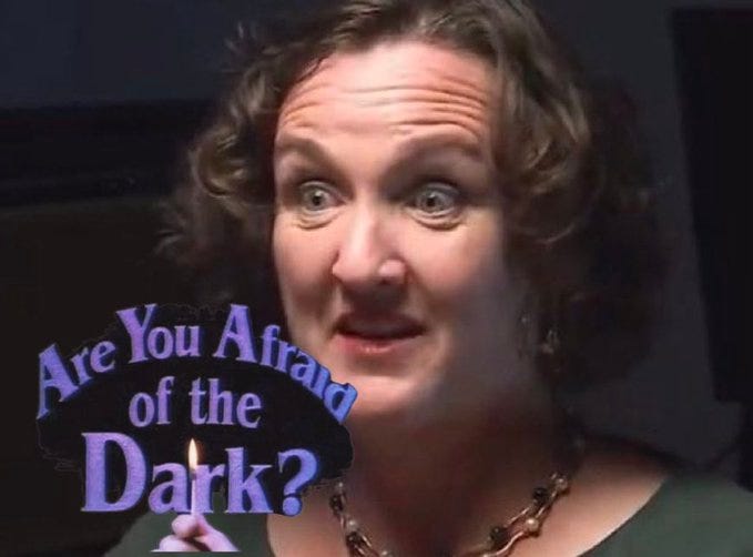 A woman with curly brown hair and wide surprised eyes holds a purple sign reading Are You Afraid of the Dark in white letters, wearing a green top and gold necklace, set against a dark background.