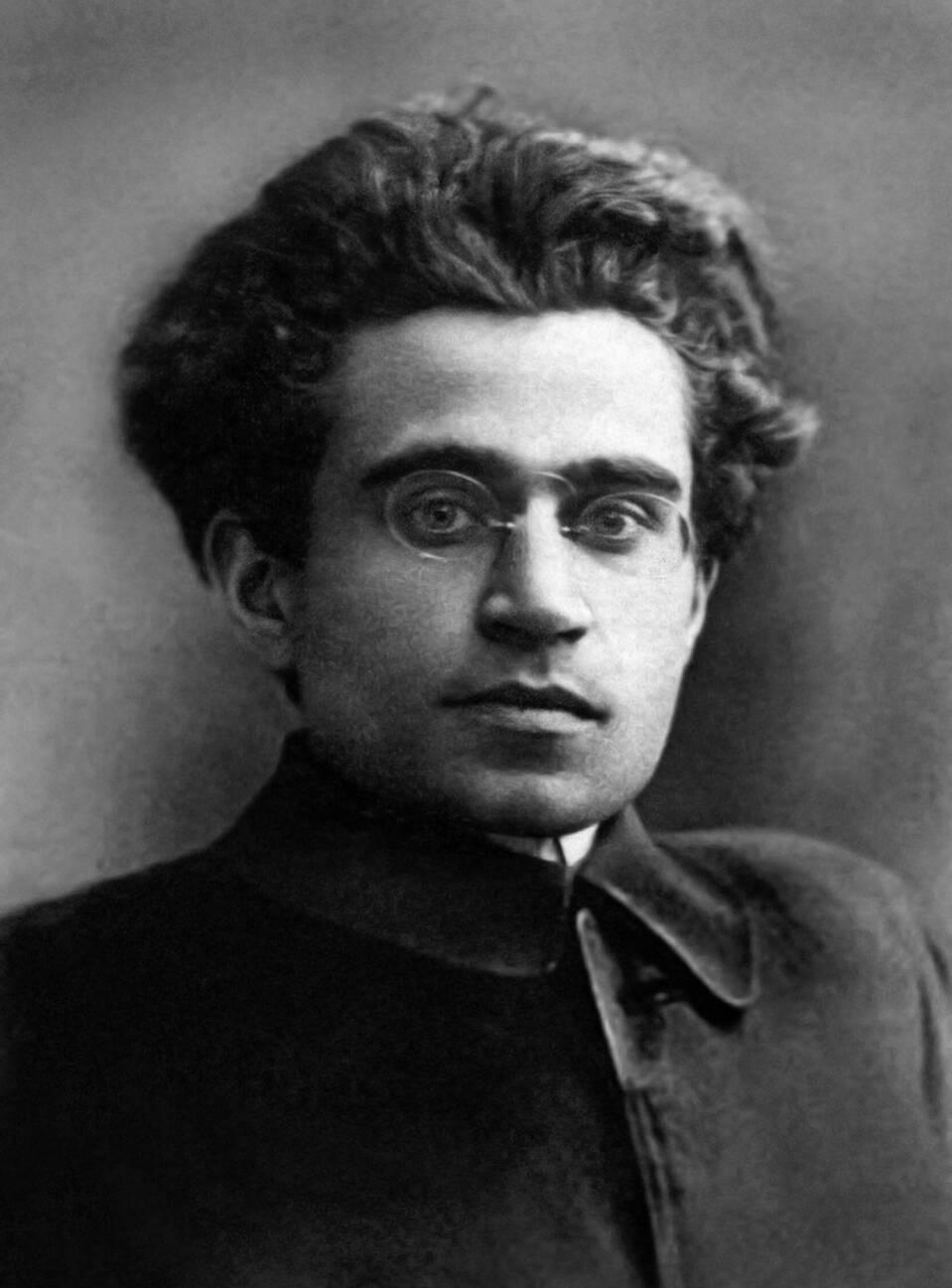 Black and white portrait photograph of Antonio Gramsci, a young man with curly hair and glasses, wearing a dark jacket.