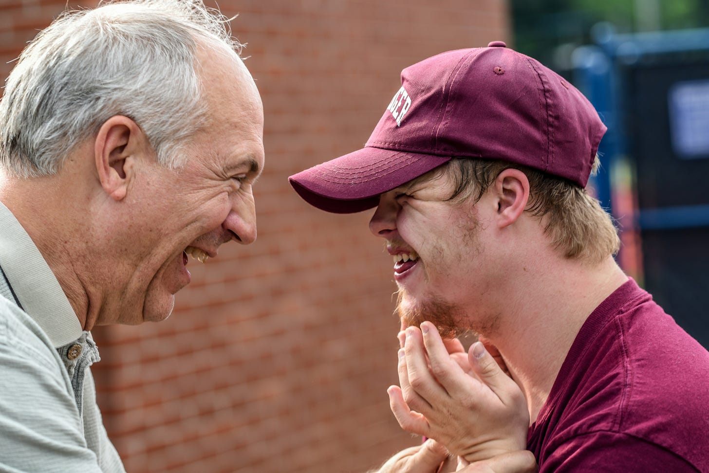Older balding grey haired man in a greenish-grey Banlon shirt with a smile of delight on his face.  A man in his teens or twenties wearing a maroon ball cap and maroon t-shirt is facing the older man.  The young man is standing so close to the older man that the brim of his ball cap is about 3 inches from the older man’s nose.  The young man, who has Down Syndrome, has both of his hands up near his reddish brown goatee. His eyes are squinted in delight and his mouth is slightly open, showing a bit of his tongue coming out from under his front teeth.  