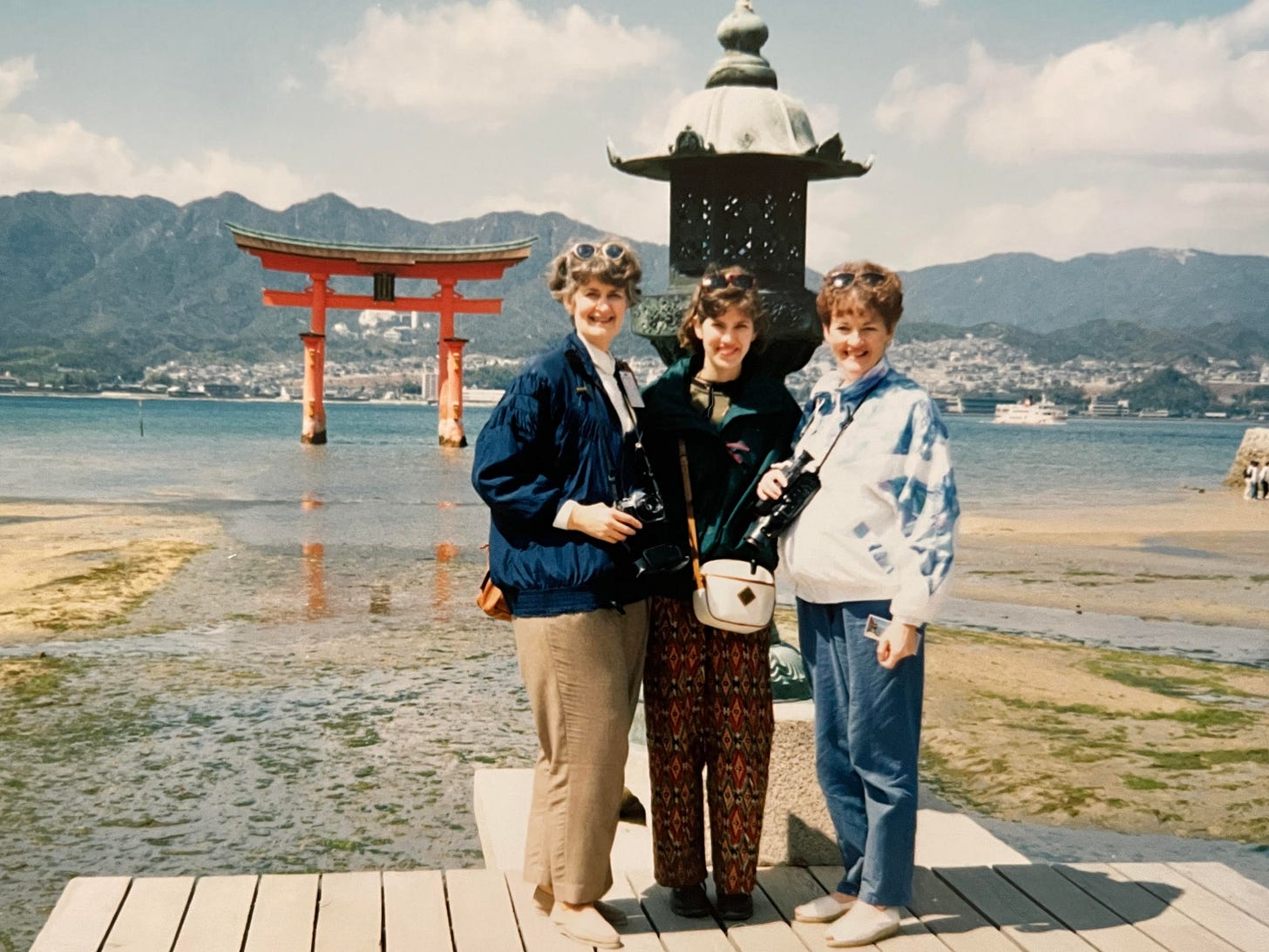 Two middle aged women and one young woman dressed in jackets and slacks, and sunglasses propped on the crowns of their heads stand in front of an 8-foot-tall (-ish) metal lantern, behind which is a huge vermilion Japanese torii gate behind which is a blue-green sea behind which are buildings and mountains and puffy clouds in a partially blue sky.