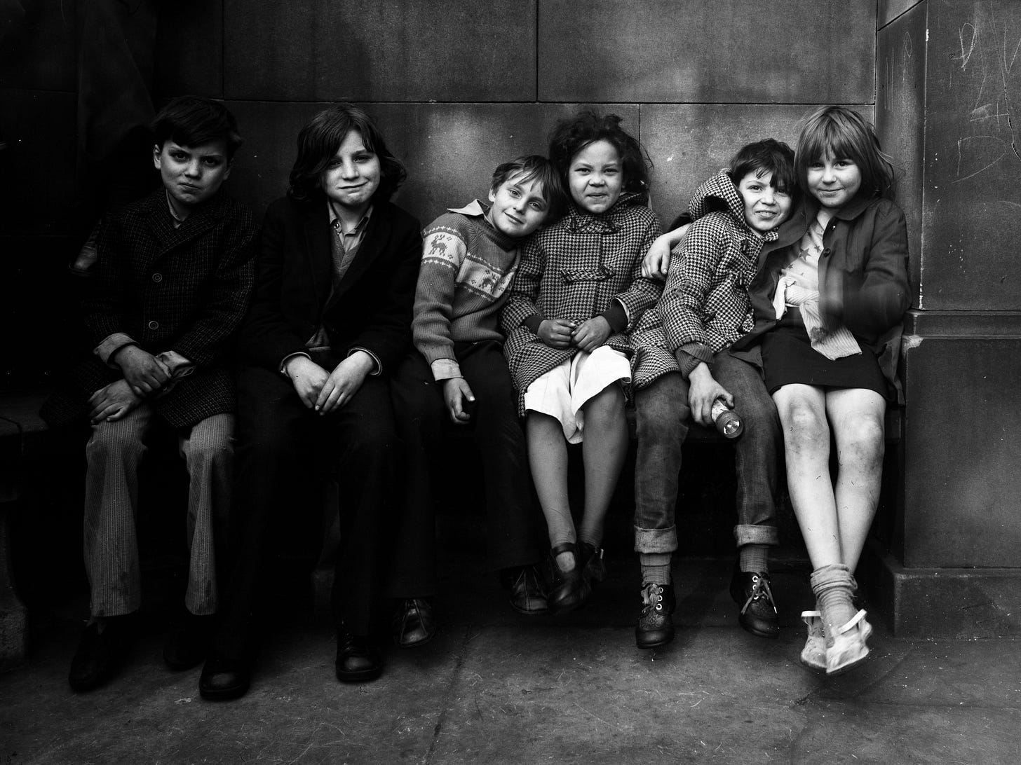 A group of six children sat on a bench smiling at the camera, with a concrete wall behind them A group of six children sat on a bench smiling at the camera, with a concrete wall behind them