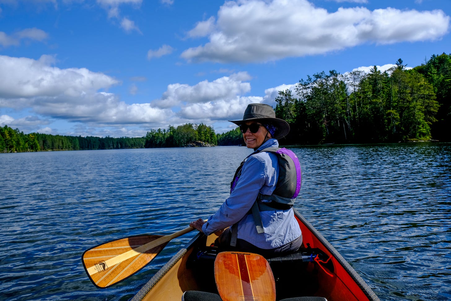 Three photographs. Left is of a clear bright sunshine-lit stretch of water bordered by tall green trees. The sun reflects off ripples on the water. In the foreground, Vicki holds her paddle at the ready while sitting in the front of her canoe. She wears a large smile.  Center photo is of a leaf littered path, tall green leafy trees on either side. In the distance, Vicki carries her canoe over her head while portaging from one stretch of water to another. Right photo shows a large stretch of water, large trees on the horizon. The sky is lit with a rising or setting sun, the clouds pink and purplish grey. The water reflects this glorious view.