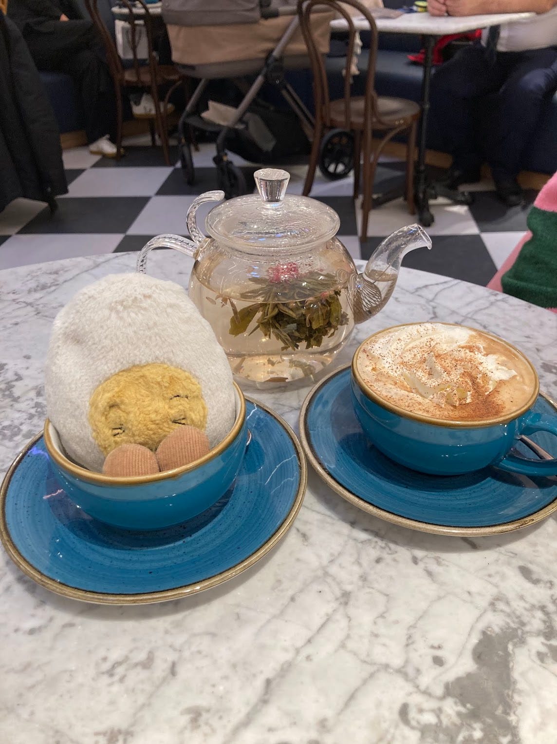 Dippy sitting in a blue tea cup, surrounded by a glass tea pot holding flower tea and a cup of mocha coffee with cream