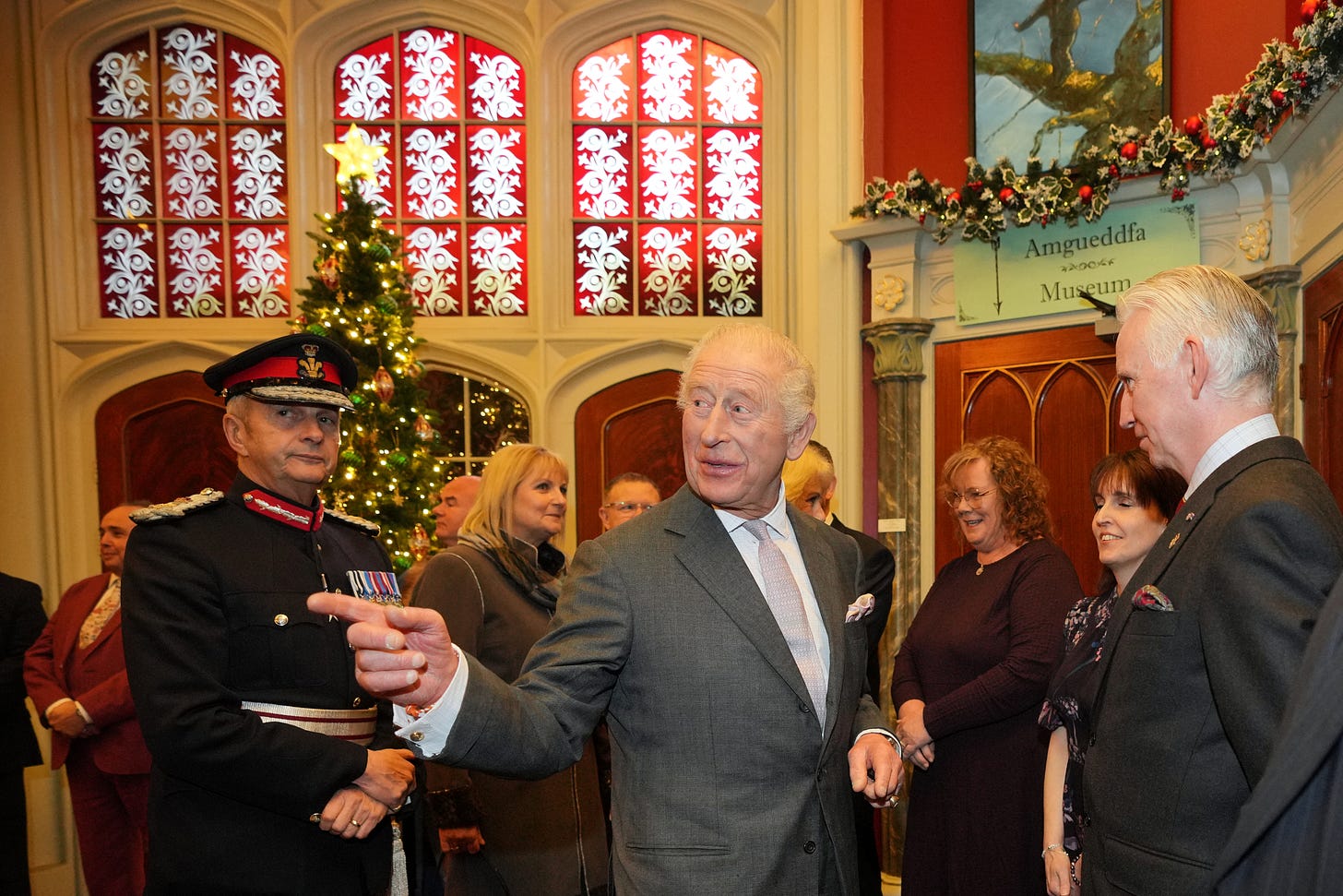 King Charles pointing at something off camera as he talks to a man in a suit inside with a Christmas tree and stained glass windows and other people in the background King Charles pointing at something off camera as he talks to a man in a suit inside with a Christmas tree and stained glass windows and other people in the background