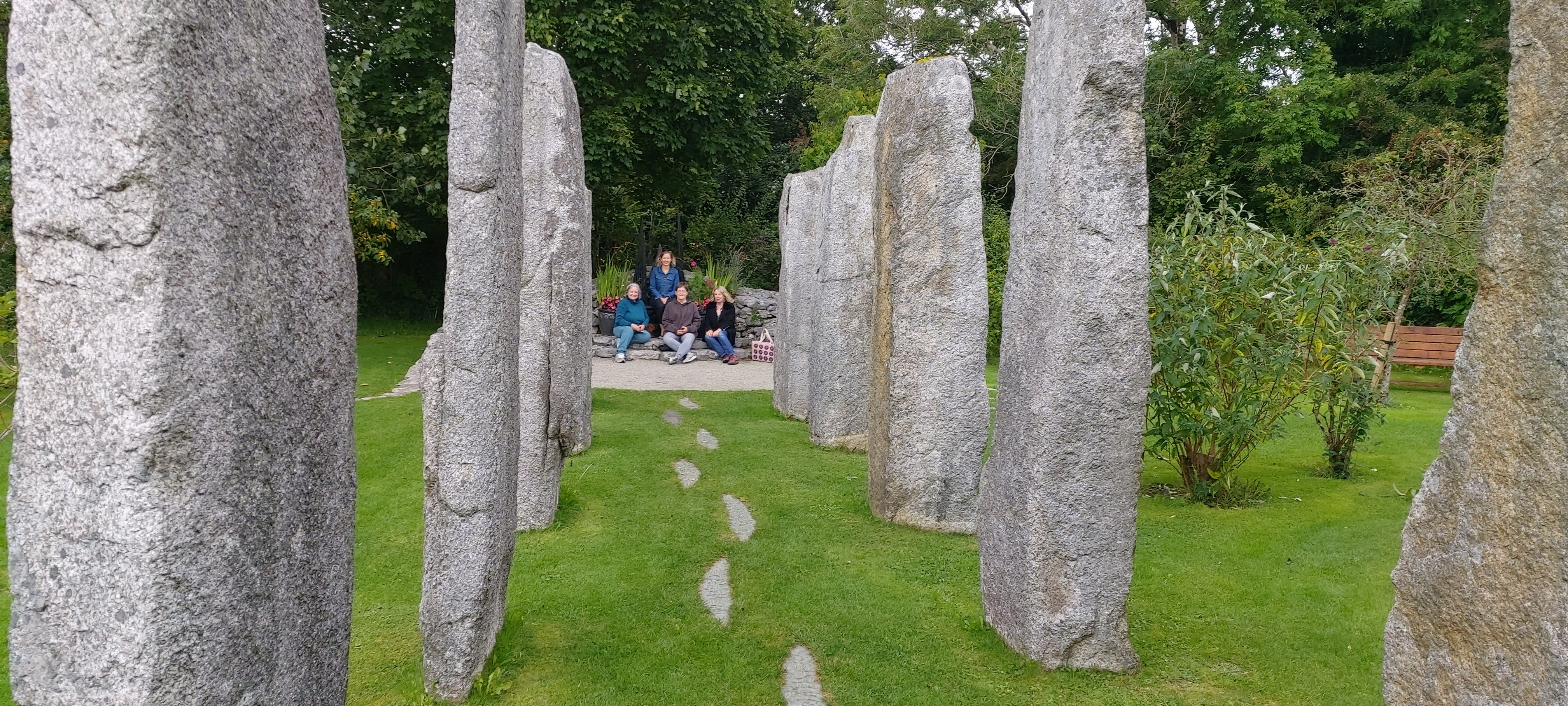 A ceremonial pathway between two stone rows leading through lush gardens with a group of 4 seated women glimpsed at the end of the passage.