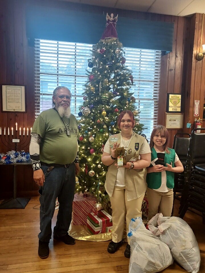 A festive indoor scene with a decorated Christmas tree topped by an angel and surrounded by wrapped presents. In front stand a bearded man in a green “U.S. ARMY” T‑shirt and two girls wearing scout uniforms—one in a tan vest with badges holding small items, the other in a green vest—next to several large filled bags. The room has wood-paneled walls, a large window with blinds behind the tree, framed certificates, a nine‑candle candelabrum resembling a menorah on the left, and stacked chairs on the right. Also, Three people pose indoors in front of a decorated Christmas tree: two youths wearing Girl Scouts uniform vests (one green, one tan) and an adult in a festive holiday vest, hat, denim skirt, and cowboy boots. The room has wood-paneled walls and a large window with blinds behind the tree. To the left is a table with a menorah and blue-and-white tinsel, while framed certificates hang on the walls and stacked chairs sit to the right on a hardwood floor. 