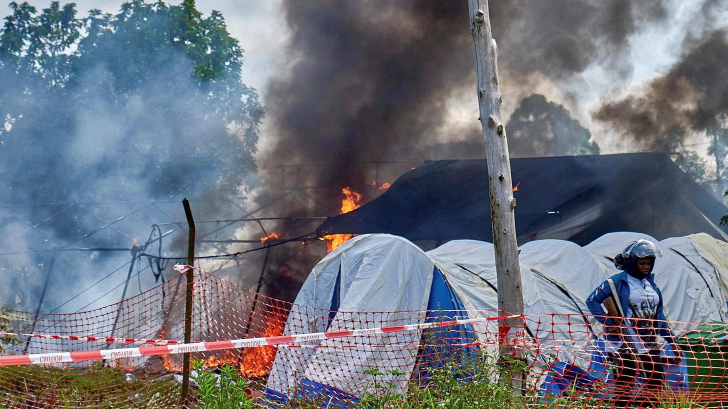 Fire and smoke at the of burnt tents on the grounds of Rwampara General Hospital. A police officer with a gun can be seen to the right of the shot in front of other makeshift tents.