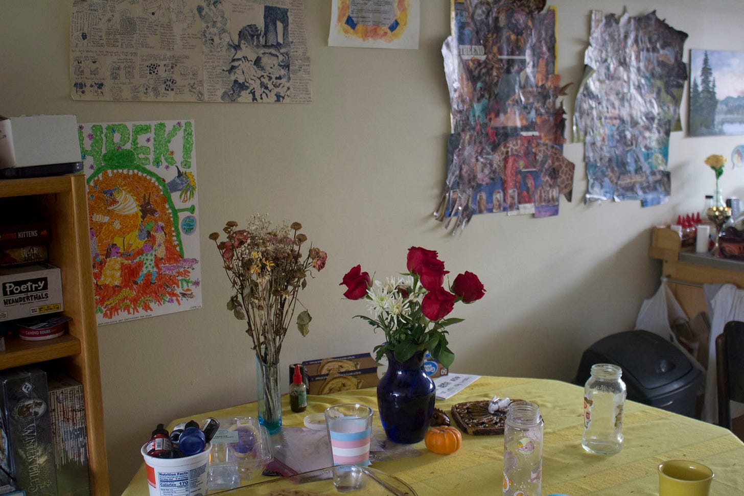 a photo of a table with flowers and drinking glasses on it. there are many drawings pinned to the wall