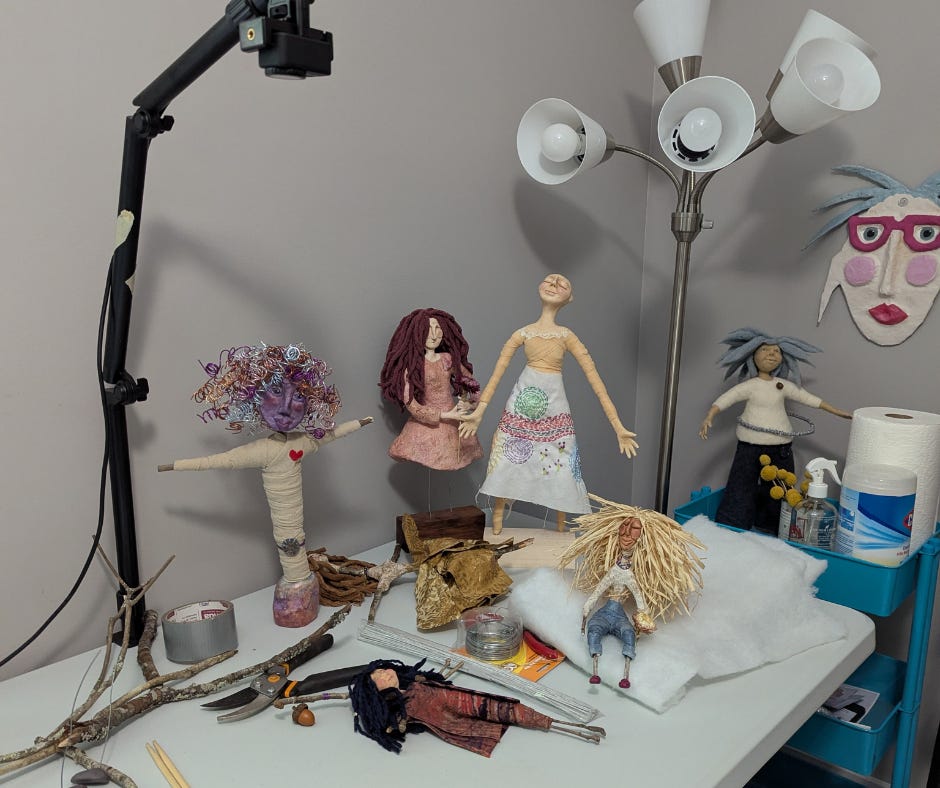 A studio table filled with handmade spirit dolls, sticks, wire, fabric, tools, and works in progress, showing a creative practice in motion. A studio table filled with handmade spirit dolls, sticks, wire, fabric, tools, and works in progress, showing a creative practice in motion.