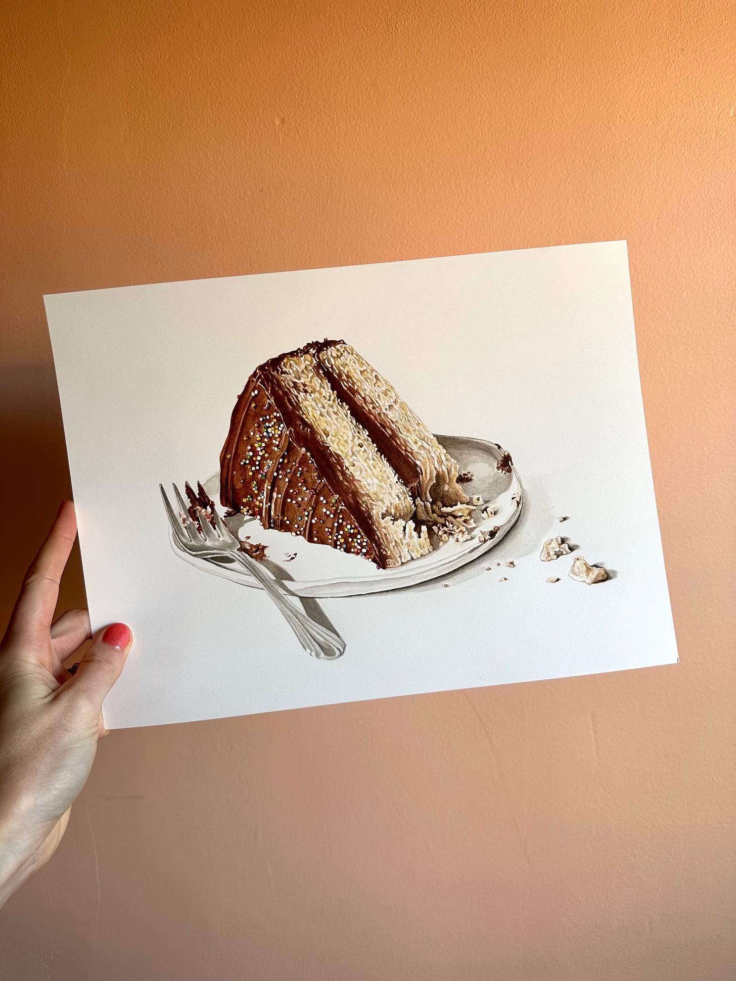 A hand holds up a painting of a slice of vanilla cake with chocolate frosting and little colorful sprinkles. A fork with crumbs sit on a small white plate. The wall behind the painting is a light peach color. 