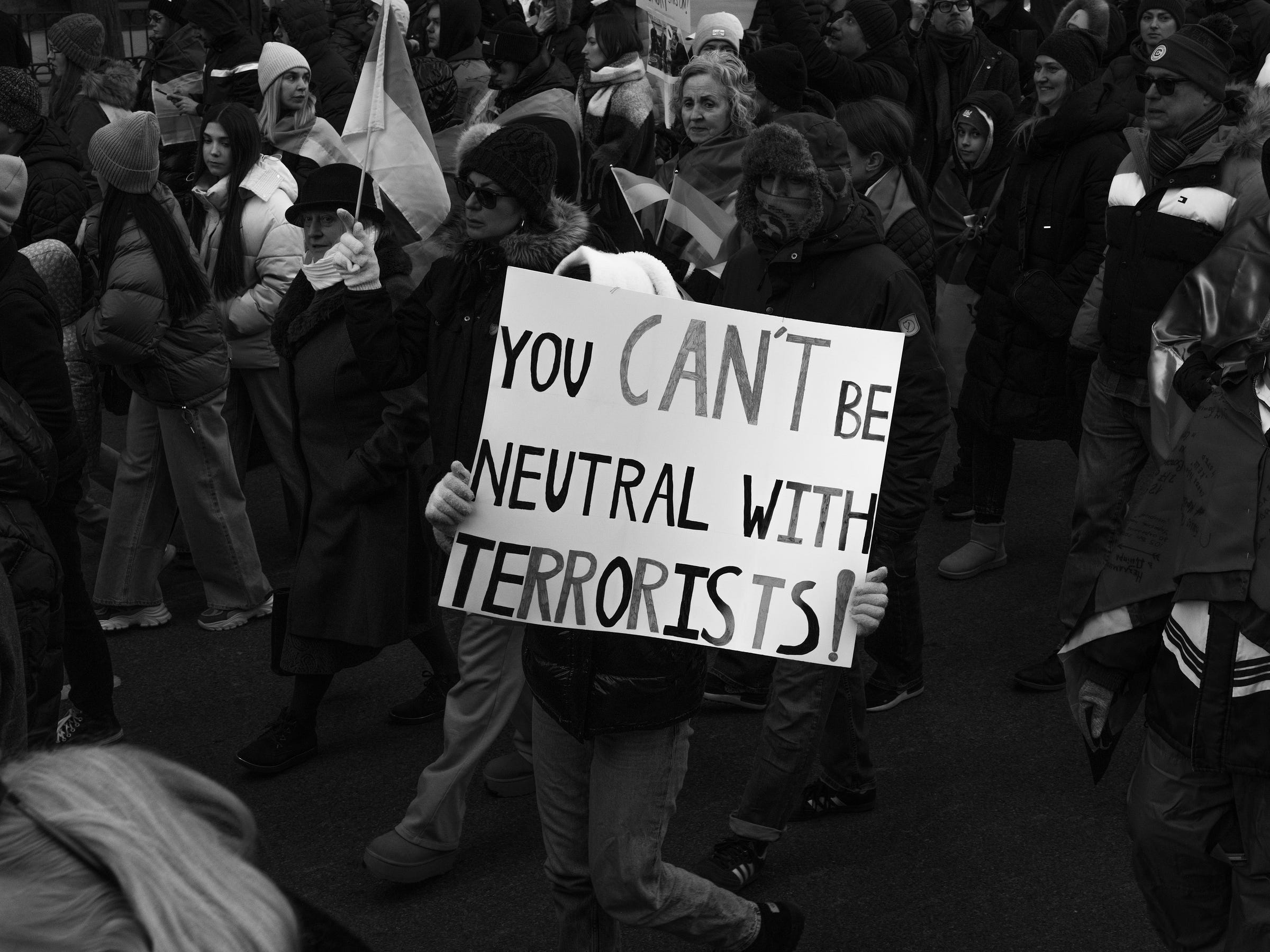 A protester holds a sign reading "YOU CAN'T BE NEUTRAL WITH TERRORISTS!" while marching with a crowd. The demonstrators, bundled in winter clothing, carry Ukrainian flags and signs, showing their solidarity. Their faces display determination as they move forward together. A protester holds a sign reading "YOU CAN'T BE NEUTRAL WITH TERRORISTS!" while marching with a crowd. The demonstrators, bundled in winter clothing, carry Ukrainian flags and signs, showing their solidarity. Their faces display determination as they move forward together.