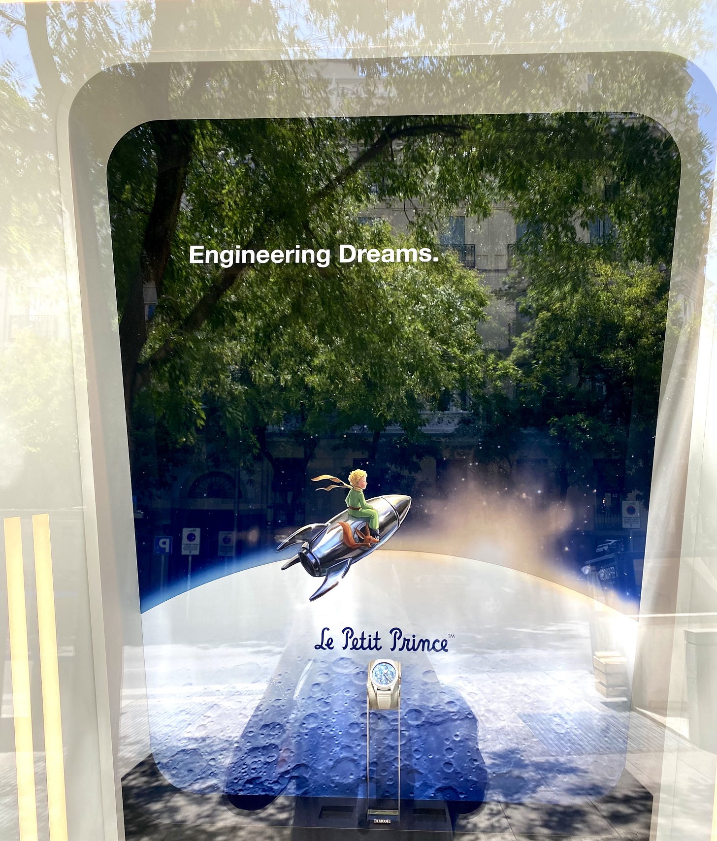 A shop window on a Madrid street. Behind the glass, a large poster shows the Little Prince riding a silver rocket above the moon, headed "Engineering Dreams.", with a pale IWC watch on a stand in front of it. Reflected in the glass, street trees and a building appear to grow out of the moon and hang in the poster's sky. A shop window on a Madrid street. Behind the glass, a large poster shows the Little Prince riding a silver rocket above the moon, headed "Engineering Dreams.", with a pale IWC watch on a stand in front of it. Reflected in the glass, street trees and a building appear to grow out of the moon and hang in the poster's sky.