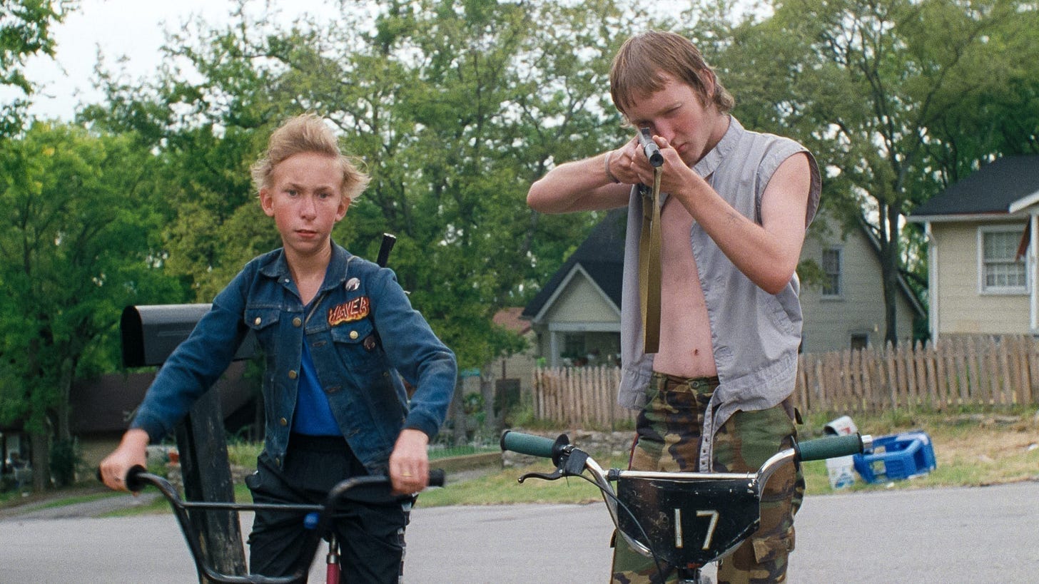 Two young southern boys on bikes, one pointing a BB gun at the camera