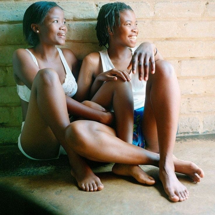 This may contain: two women sitting next to each other in front of a brick wall with their arms around one another