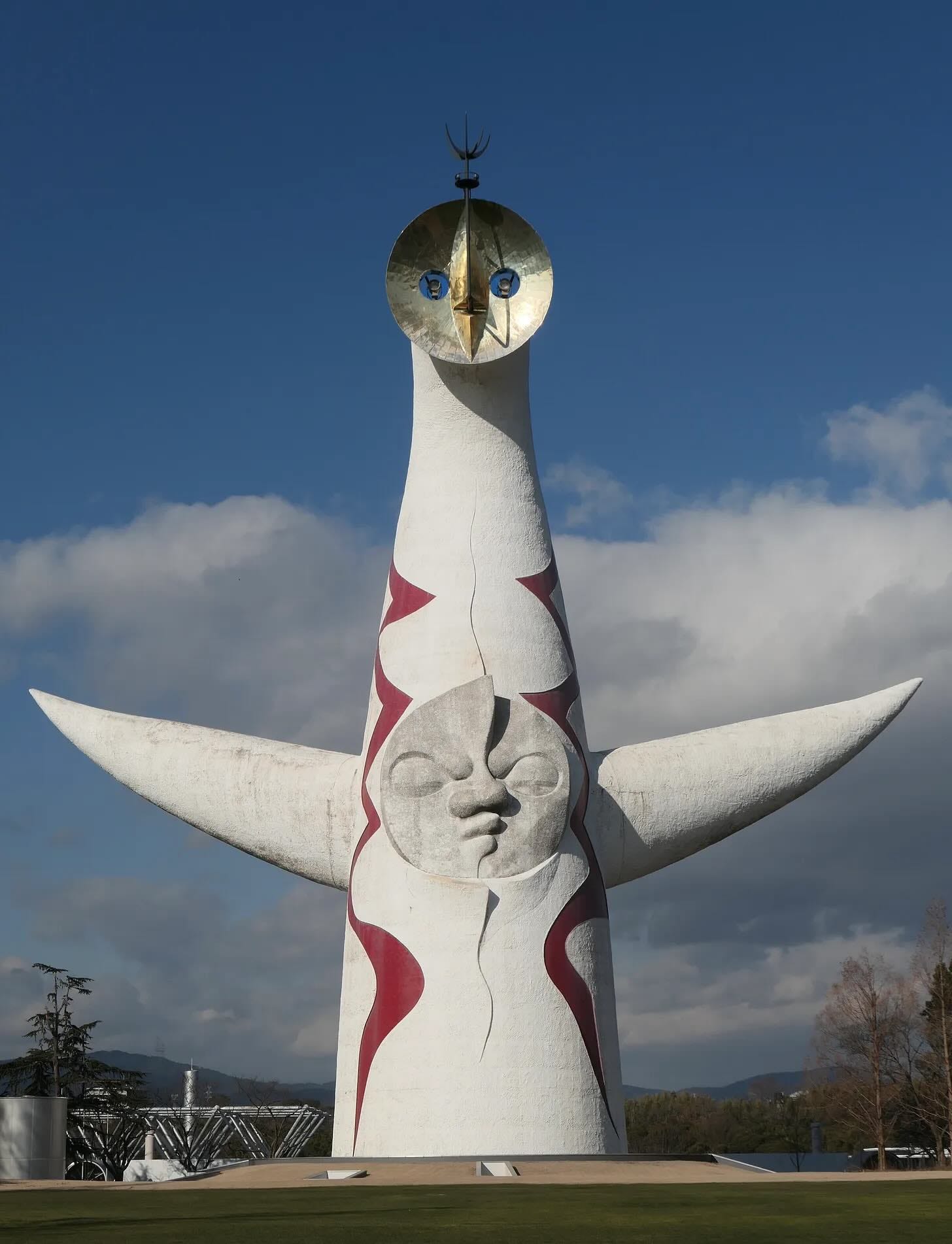 A large white sculpture in a park under a blue sky with some clouds. The sculpture is very roughly shaped like a human body, with a bottle-shaped body, 2 outstretched conical arms, and a face above the body. The body has a face in the middle, with a possibly angry expression, and 2 red zigzag patterns on either side of the face. The face at the top of the sculpture is a silver disk, with 2 holes for eyes, and a vertical pointed oval that represents the nose and mouth.