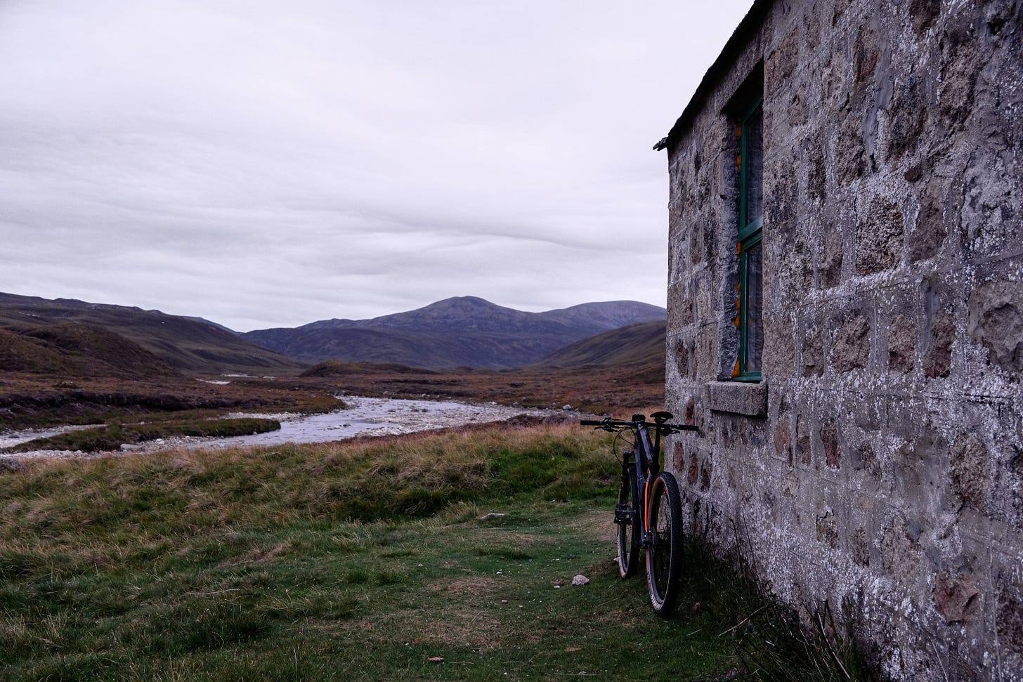 A bicycle rests against the outside wall of a small stone bothy in the Highlands. Beyond, open hills and a winding river create a sense of vastness and remoteness, contrasting with the intimacy of the shelter.