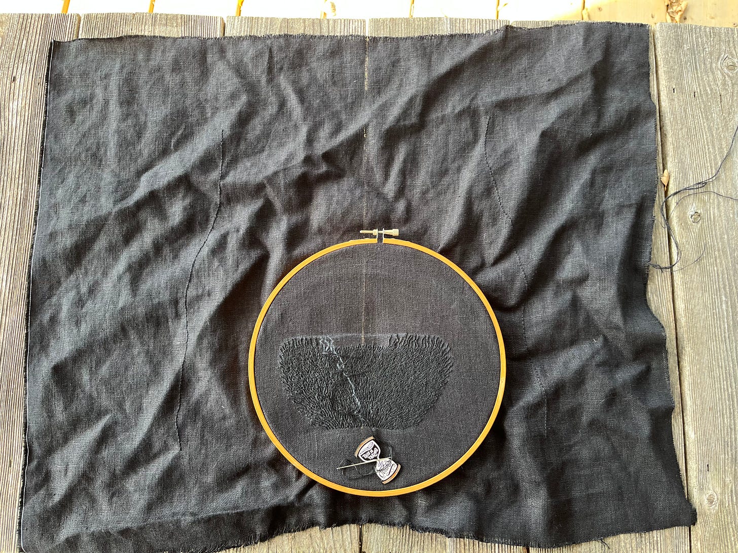 The lower torso and hips of a woman are outlined in black thread on wrinkled black linen. The public hair is in an embroidery hoop and being stitched in black thread. An hourglass-shaped needle minder that reads “this is taking forever” is attached to the cloth.  