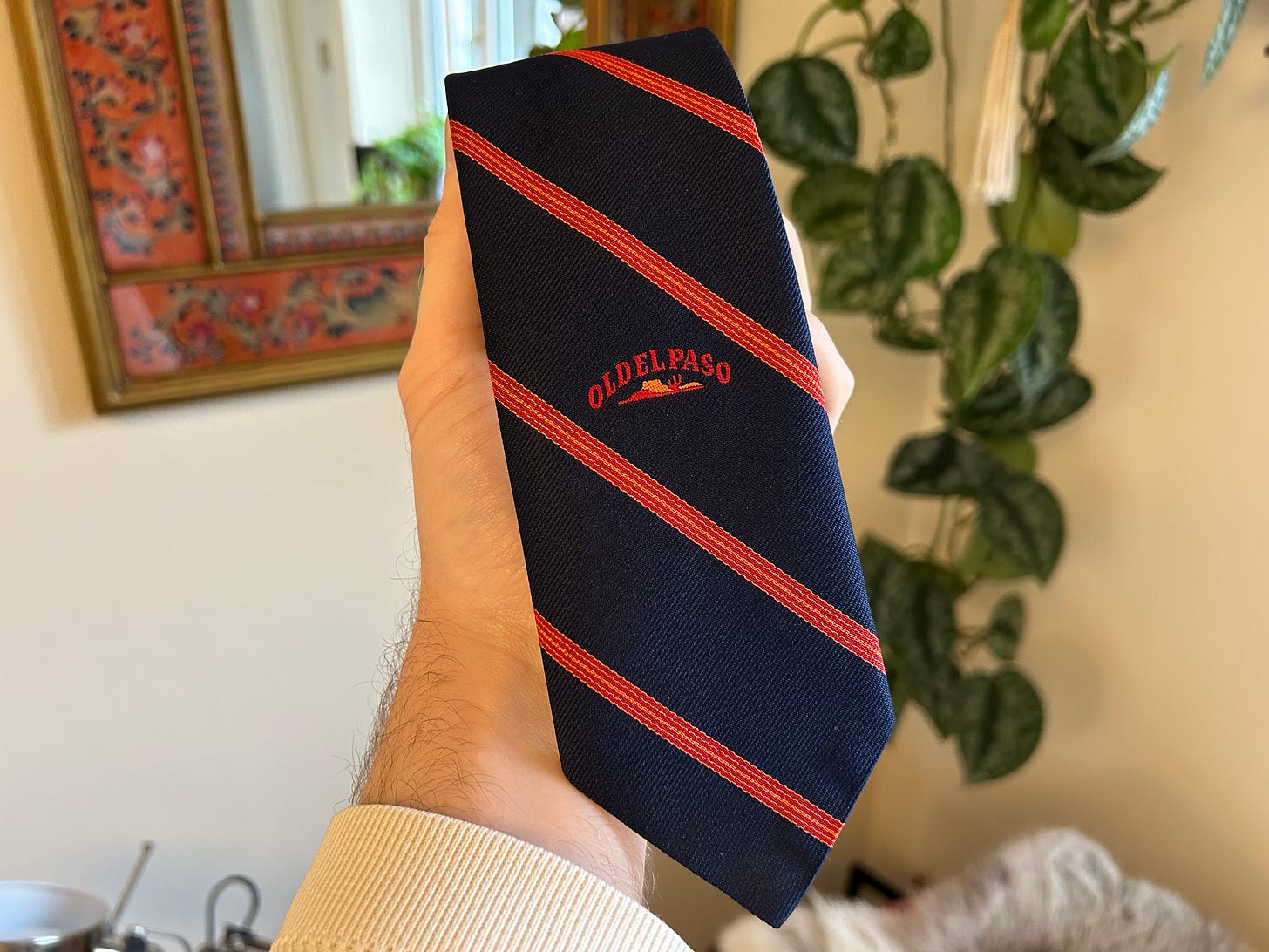 A masculine hand holds a blue tie with widely spaced orange, diagonal stripes and the "Old El Paso" logo is embroidered on it. The tie is rolled up. The background is the corner of a magnolia room with a nice hanging plant and the edge of a mirror but the focus of the image is the tie.