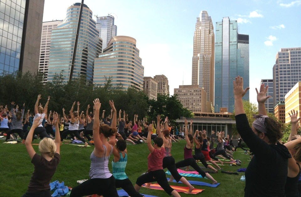 A crowd of people do yoga on a lawn in downtown Minneapolis, with skyscrapers surrounding them.