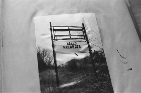 1) A black & white photograph of a photograph, which contains a large sign on the side of the road reading: "HELLO STRANGER" in capital letters. 2) A scattered pile of multi-coloured books overlayed with sheer, green pattered cloth. 3) A black & white photograph of café chairs & tables crammed against a wall. 4) Scattered, multi-coloured books. 5) A black & white photograph of a wooden deck in an alleyway. Lights are strung across both the deck & alleyway. 6) An ink drawing of a map of a fantasy land. 7) A black & white photograph of two chairs & a table next to a window. A neon sign reading "coffee" is above them. 8) A black, mauve, and dark green watercolour painting of a bare forest. Beneath the painting are the words "magic forest" in calligraphy. 9) A black & white photograph of the roof of an ornate building, mirroring itself at the top & bottom of the image.