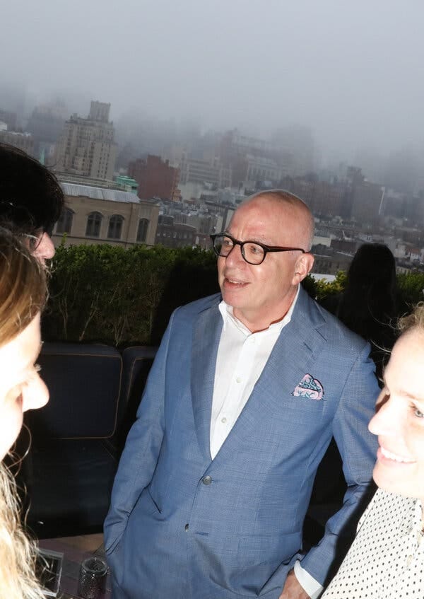 A person in a blue suit, white shirt, and glasses speaks to others, a patterned pocket square visible. A hazy city skyline is in the background. A person in a blue suit, white shirt, and glasses speaks to others, a patterned pocket square visible. A hazy city skyline is in the background.
