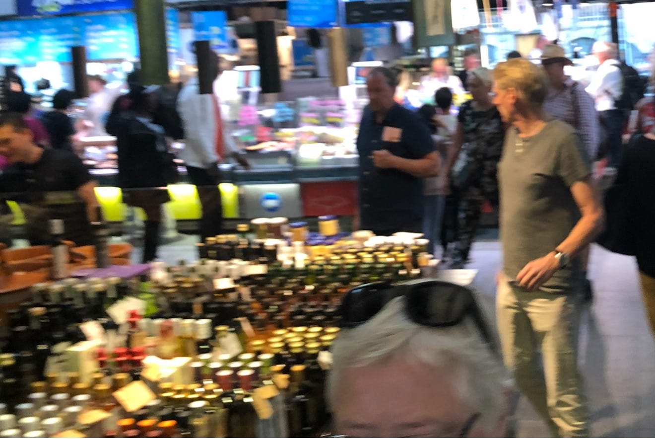 Blurred indoor market scene with a crowded table of bottles in the foreground and shoppers moving between fresh-food stalls.