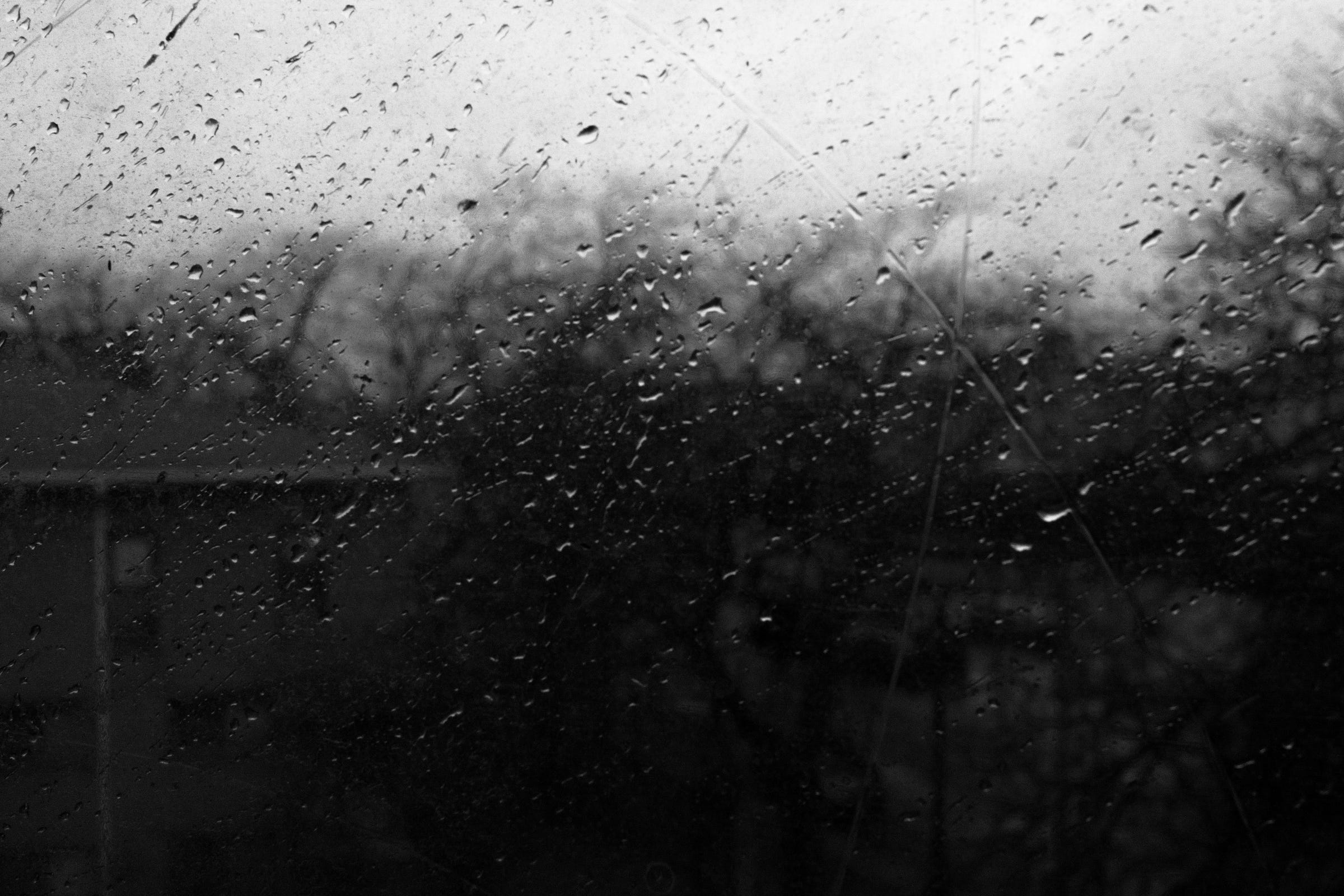 Rain droplets and streaks on a scratched window, with a blurred view of trees and buildings in the background on a gloomy day. Rain droplets and streaks on a scratched window, with a blurred view of trees and buildings in the background on a gloomy day.