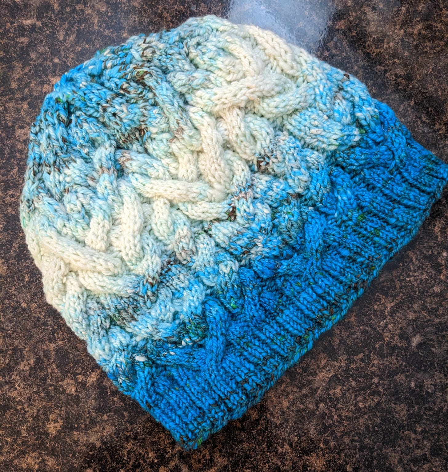 A photo of a hand-knit cabled hat in a gradient that runs from dark turquoise to cream and back to turquoise.