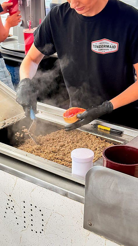 View of line outside of the door. Employee scooping ground beef onto a bun. Closeup of the menu on an old school board.