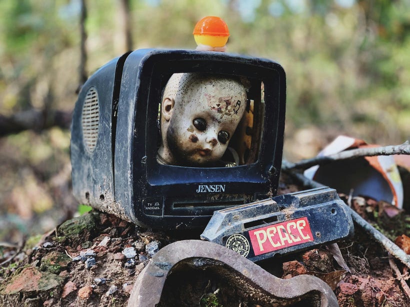 An ancient black Jensen television frame sits broken and dusty on a small heap of dirt, rocks, and other junk. Where the screen used to be is a dirty, hairless doll's head. In front of this is an old metal part of some kind, with a red sticker on it that says "PEACE" in white letters. The background is blurred but it looks like we're in a wooded area on a sunny day. An ancient black Jensen television frame sits broken and dusty on a small heap of dirt, rocks, and other junk. Where the screen used to be is a dirty, hairless doll's head. In front of this is an old metal part of some kind, with a red sticker on it that says "PEACE" in white letters. The background is blurred but it looks like we're in a wooded area on a sunny day.