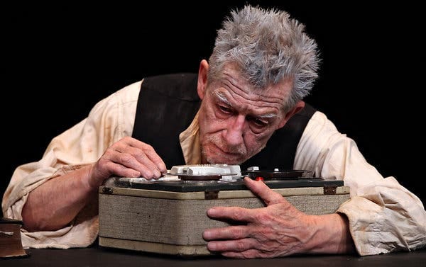 John Hurt, with spiky grey/white hear and wearing a black waistcoat over a white shirt, leans over a large tape player/recorder.