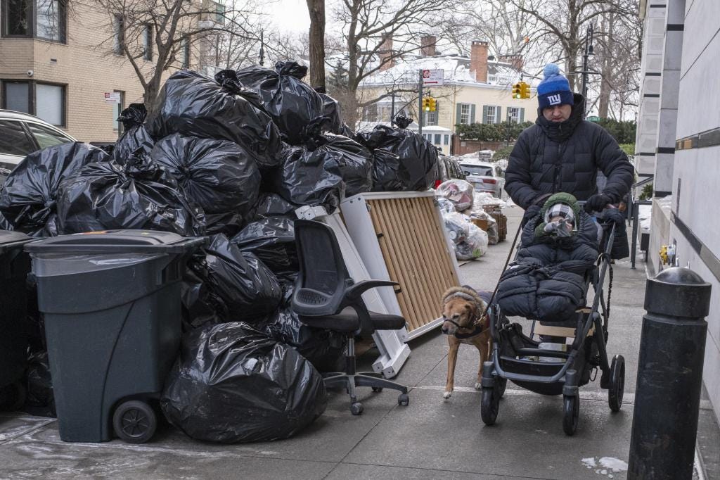 A man in a blue hat pushes a child in a stroller past large piles of black trash bags and a dog. A man in a blue hat pushes a child in a stroller past large piles of black trash bags and a dog.