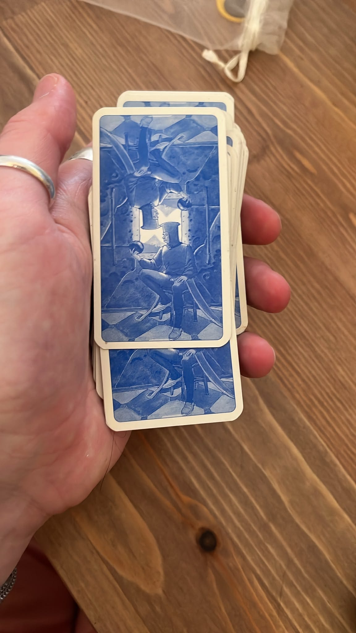 A hand holds a small tarot deck with its blue illustrated card backs visible, showing two seated figures in a surreal interior. A hand holds a small tarot deck with its blue illustrated card backs visible, showing two seated figures in a surreal interior.