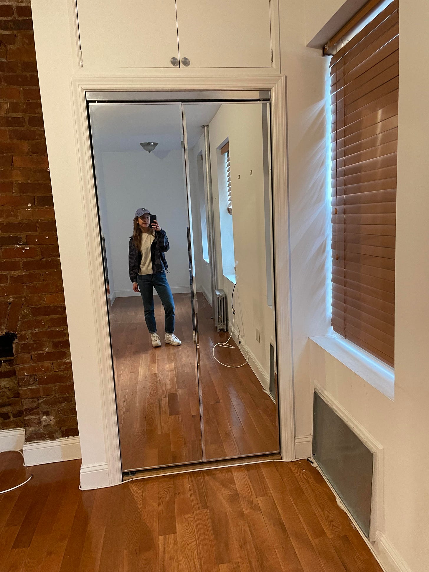 Sara takes a full-length mirror selfie in the sliding closet door of her apartment in Kips Bay, with hardwood floors, exposed brick, and a window covered with blinds. Sara takes a full-length mirror selfie in the sliding closet door of her apartment in Kips Bay, with hardwood floors, exposed brick, and a window covered with blinds.