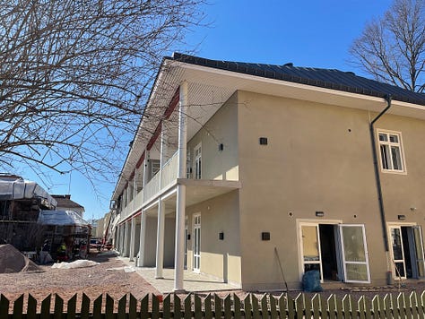 A two story building in beige with a dark grey roof form 2 angles. A group of shirtless older men standing on their balcony looking out into my balcony