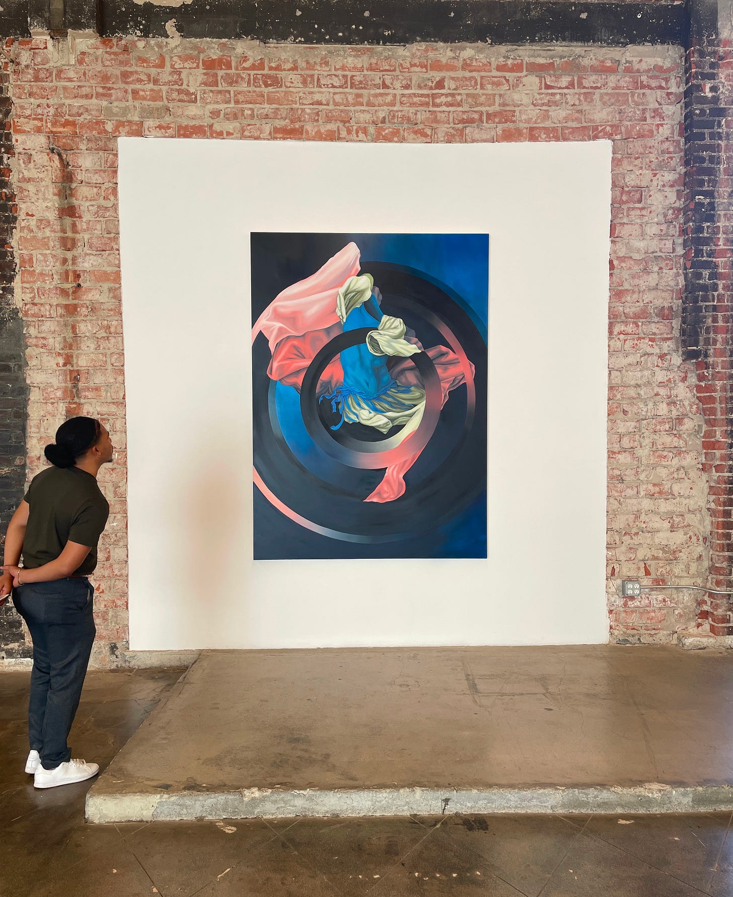 Man in dark green shirt, dark gray slacks, and white sneakers standing with his arms behind his back. He is gazing upward at a large oil painting with swirles of pink, blue, and cream garments woven throughout a black target on a blue background Man in dark green shirt, dark gray slacks, and white sneakers standing with his arms behind his back. He is gazing upward at a large oil painting with swirles of pink, blue, and cream garments woven throughout a black target on a blue background