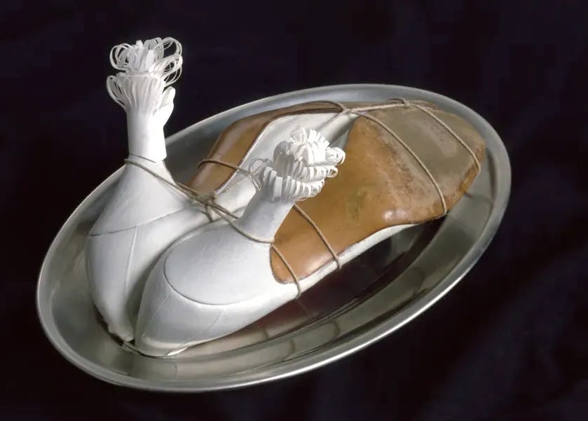 A pair of women’s high-heeled shoes arranged on a silver platter like a trussed roast, bound with string and topped with decorative paper frills on the heels, referencing food presentation.