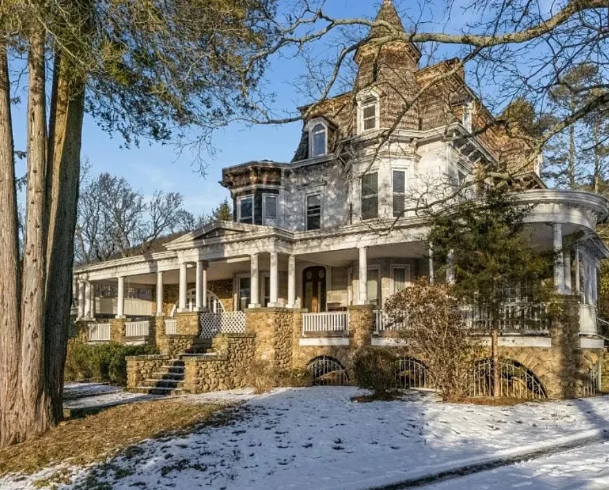 A large Victorian house with a turret and wide wraparound porch supported by white columns sits on a wooded property with patches of snow on the ground. Stone steps lead up to the porch, and tall trees frame the historic building, which was once the centerpiece of the Goodland Country Club nudist resort in New Jersey.