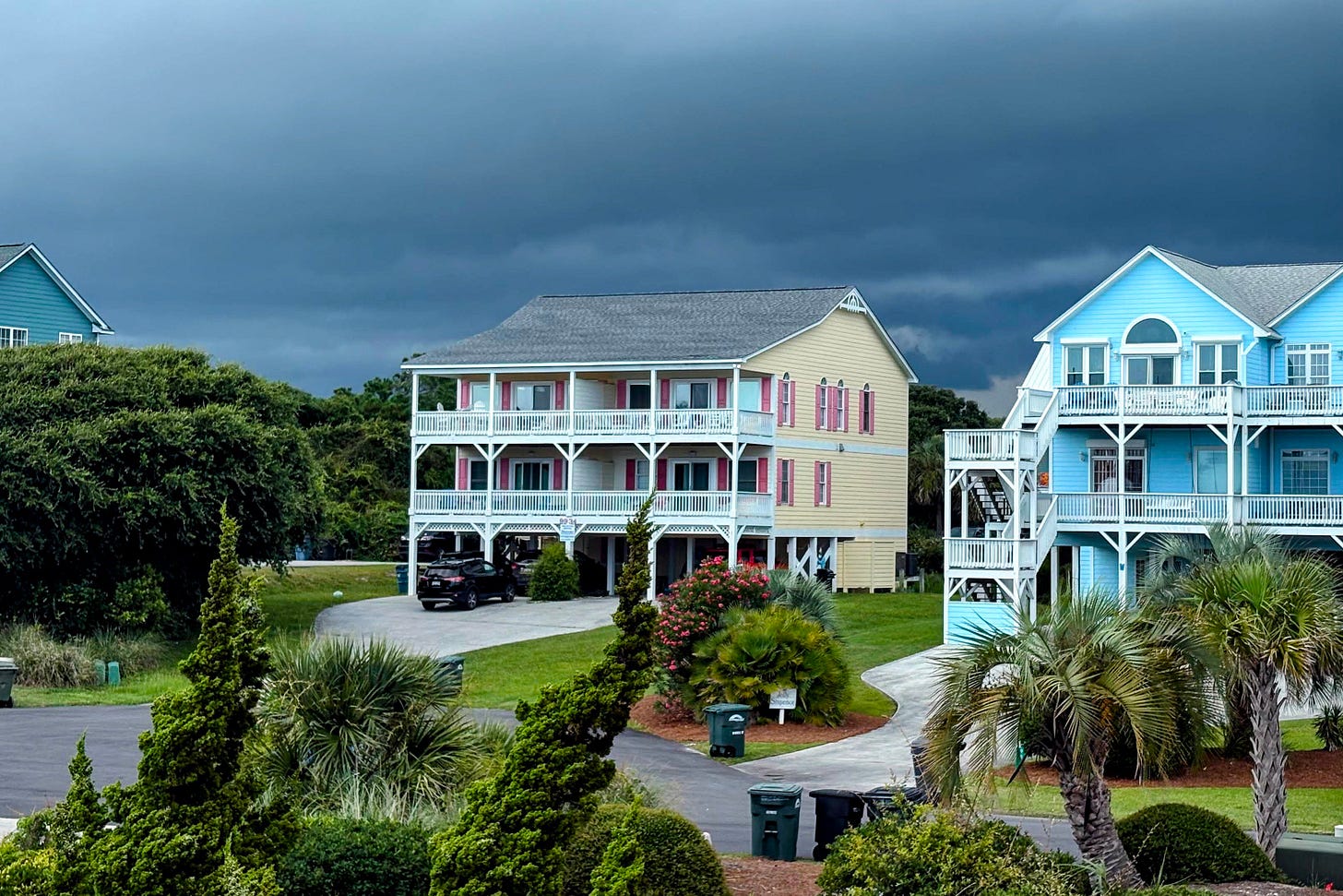 Large pastel-colored beach houses stand beneath dramatic dark storm clouds, surrounded by palm trees and manicured lawns in a coastal neighborhood.