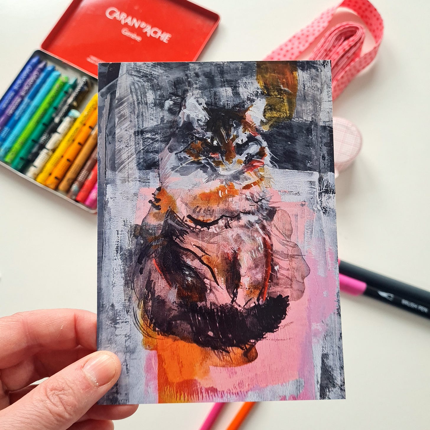Hand holding up a greeting card with a cat painting on the front. In the background are art materials. Hand holding up a greeting card with a cat painting on the front. In the background are art materials.