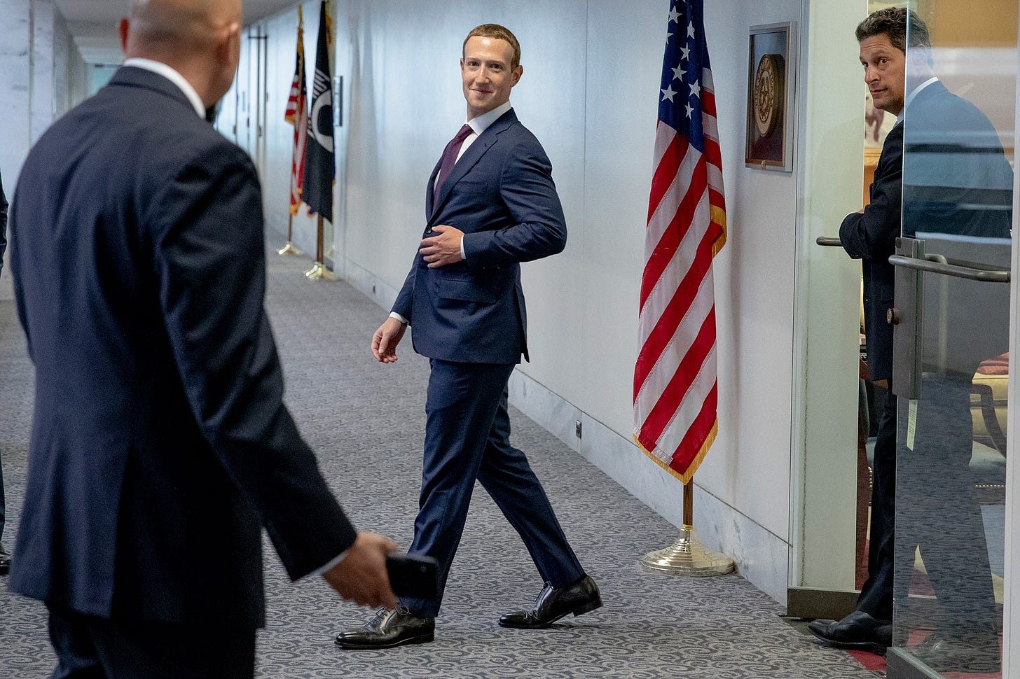 Mark Zuckerberg walks out of a room into a government office hallway next to an American flag. Mark Zuckerberg walks out of a room into a government office hallway next to an American flag.