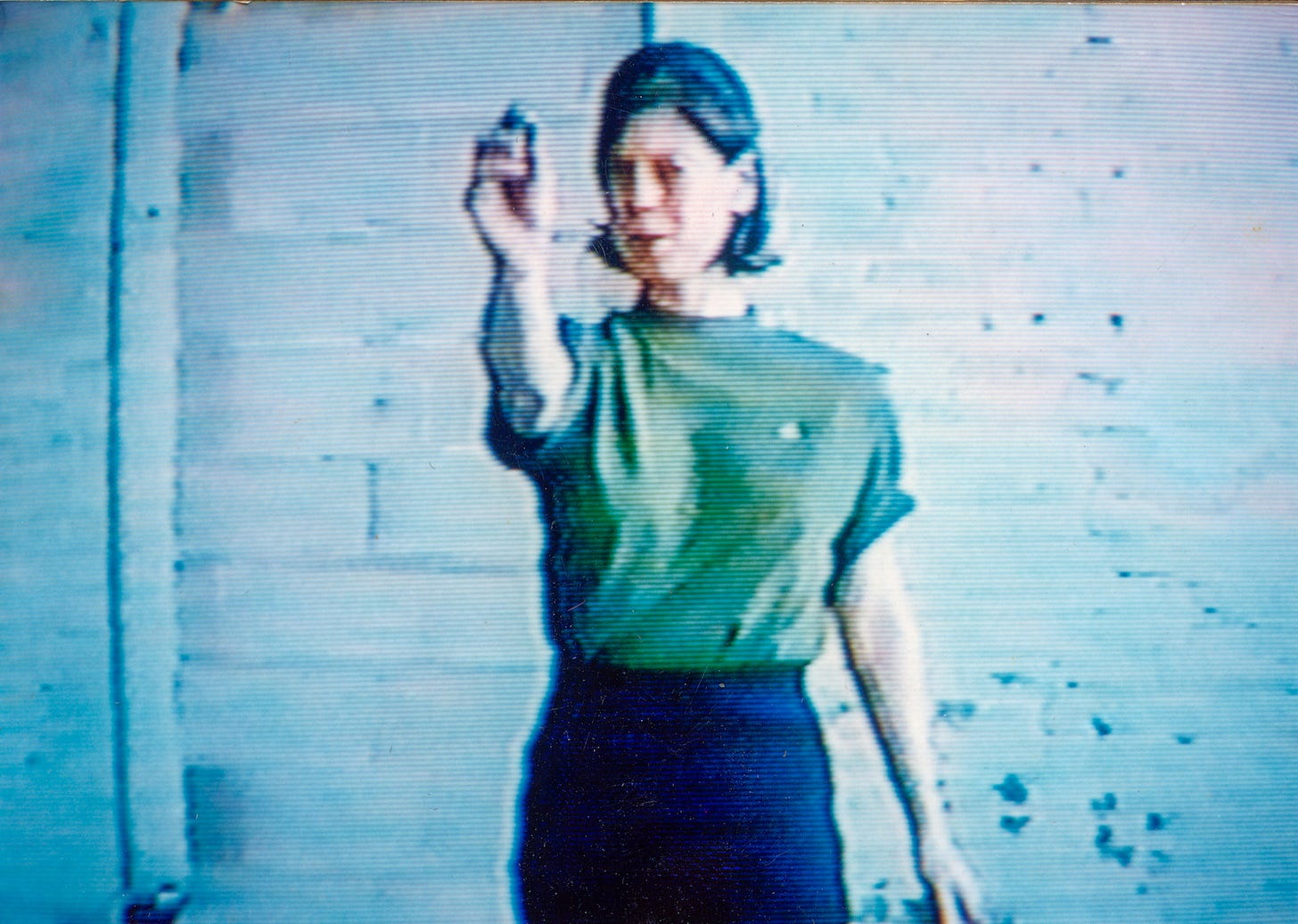 Lo-res video still of the artist, Mary Addison Hackett standing in an empty room facing the camera. Her right hand is extended as she prepares to throw a dart. She’s wearing a short-sleeve green shirt and black pants. Lo-res video still of the artist, Mary Addison Hackett standing in an empty room facing the camera. Her right hand is extended as she prepares to throw a dart. She’s wearing a short-sleeve green shirt and black pants.