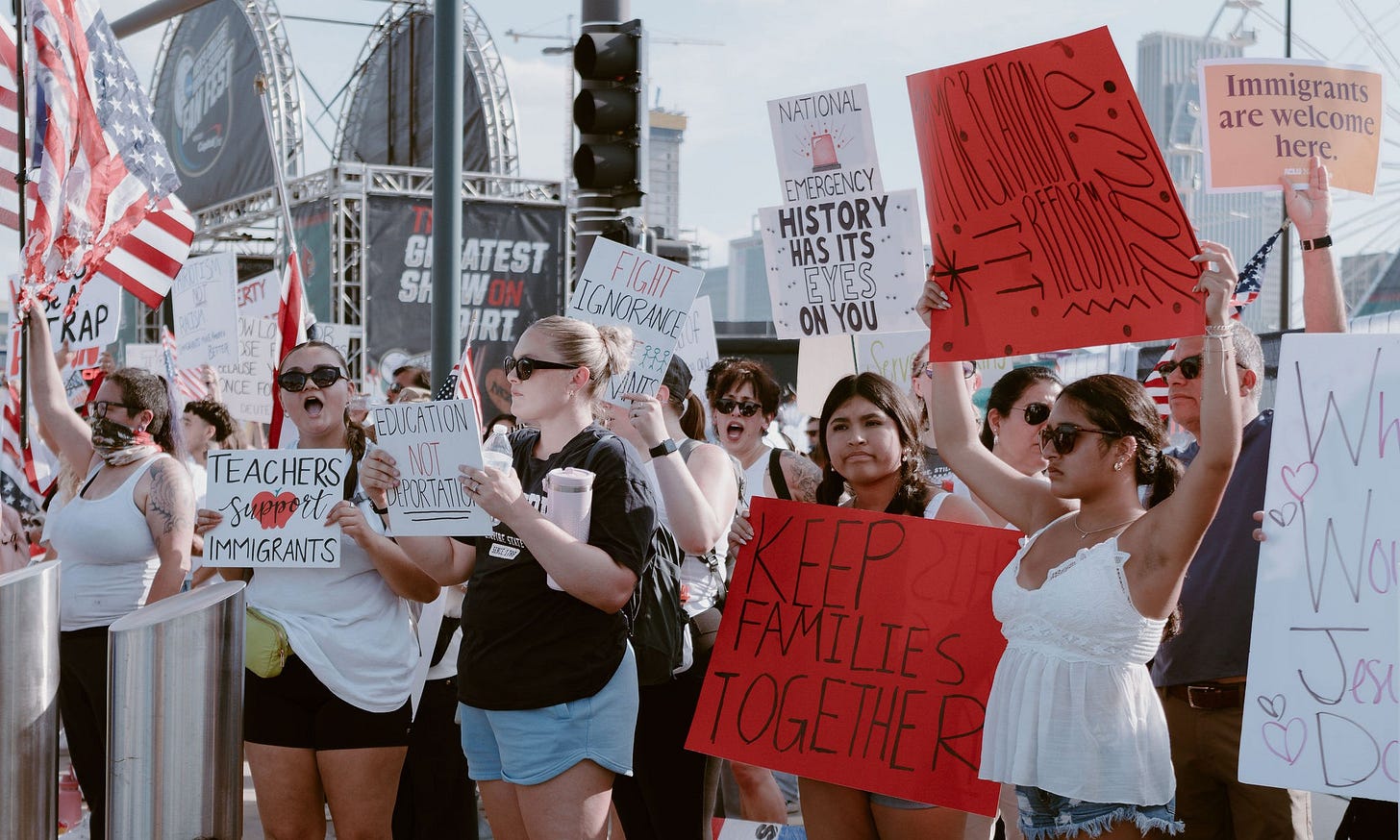 A diverse group of peaceful protesters stands together outside the College World Series in Omaha, holding signs with messages such as “Keep Families Together,” “Teachers Support Immigrants,” “Education Not Deportation,” and “Immigrants Are Welcome Here.” An American flag is visible among the crowd, and downtown Omaha buildings can be seen in the background. The image captures a powerful moment of community solidarity and advocacy in response ICE actions that impacted local families. A diverse group of peaceful protesters stands together outside the College World Series in Omaha, holding signs with messages such as “Keep Families Together,” “Teachers Support Immigrants,” “Education Not Deportation,” and “Immigrants Are Welcome Here.” An American flag is visible among the crowd, and downtown Omaha buildings can be seen in the background. The image captures a powerful moment of community solidarity and advocacy in response ICE actions that impacted local families.